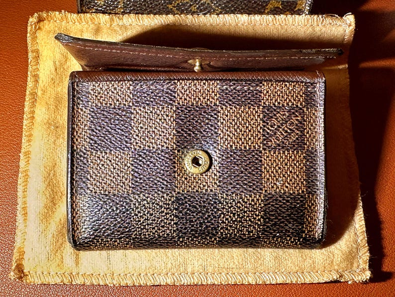 May include: A brown and black checkered wallet with a gold button clasp. The wallet is open, revealing a brown interior. The wallet is resting on a tan cloth.