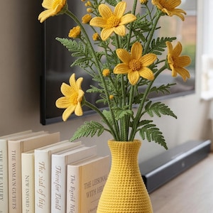 May include: A yellow crochet vase holding crochet yellow flowers and green leaves. The vase is on a wooden surface beside a row of books. The text "KNOTGARDENDIGITAL" is on the vase.