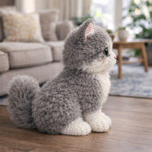 May include: A plush gray and white kitten toy with a fluffy tail, sitting on a wooden floor. The kitten has large, dark eyes and pink accents on its ears and cheeks. The toy is in a seated position, with its paws visible.