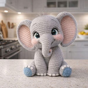 Puede incluir: Un juguete de elefante de ganchillo en posición sentada. El elefante es gris con orejas grandes de color rosa claro y mejillas rosadas. Sus pies son azules. El juguete tiene ojos grandes y expresivos y una pequeña trompa. El fondo es una cocina.