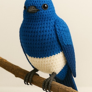 Könnte beinhalten: Ein handgefertigtes blaues Vogelplüschtier mit weißem Bauch, das auf einem braunen Ast sitzt. Der Vogel hat schwarze Augen und Füße und besteht aus einem strukturierten, gestrickten Material. Ein dekorativer Artikel.