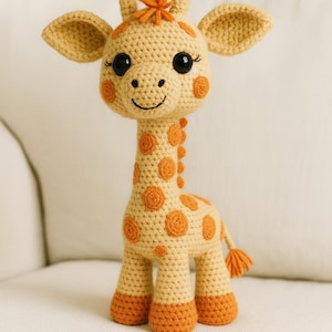 以下が含まれることがあります： 黄色とオレンジ色の手編みのキリンのおもちゃ。キリンは大きな黒い目、オレンジ色の斑点、笑顔の顔をしています。おもちゃの高さは約25cmです。
