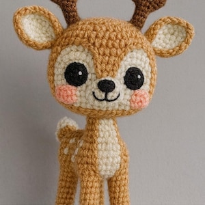 May include: A crocheted deer toy in shades of tan, cream, and brown. The deer features large black eyes, pink cheeks, and small brown antlers. The body is tan with a cream belly and white spots on the back.