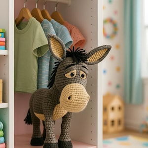 May include: A handmade, gray crochet donkey toy with a tan muzzle and ears stands in a white closet. The donkey has black eyes and a sad expression. Several children's t-shirts hang on wooden hangers above the donkey.