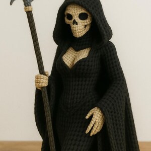 May include: Crocheted Grim Reaper figurine in a black hooded robe, holding a gray scythe with a tan handle. The skull and hands are tan. The figure is standing on a wooden surface.