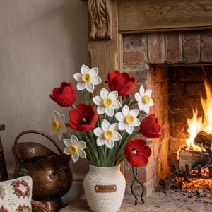 以下が含まれることがあります： 赤のチューリップと黄色の中心を持つ白い水仙の、クリーム色の花瓶に入ったかぎ針編みのブーケ。花瓶は、火の燃えるレンガの暖炉の前の石の暖炉の上にあります。銅製のバケツと編まれたバスケットが背景にあります。