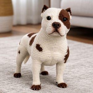 May include: A crochet dog figurine in a standing pose. The dog is mainly off-white with brown patches and accents on the ears, eyes, and paws. The dog is standing on a grey rug.