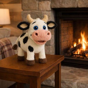 Può includere: Una statuetta di mucca all'uncinetto fatta a mano con corpo bianco, macchie nere e muso rosa. La mucca ha occhi blu, corna beige e zoccoli marroni. È esposta su un tavolo di legno davanti a un camino.