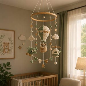 May include: A handcrafted baby mobile with a hot air balloon, aeroplane, and helicopter, all made from yarn. The mobile includes wooden stars, clouds, and a banner. The colour scheme is soft, with shades of green, beige, and white.