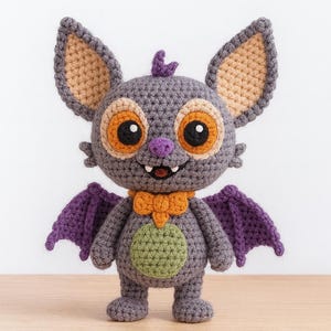 Puede incluir: Muñeca de murciélago de ganchillo hecha a mano en tonos grises, morados, naranjas y verdes. El murciélago tiene ojos grandes, una pequeña sonrisa y alas detalladas. La muñeca está sobre una superficie de madera clara.