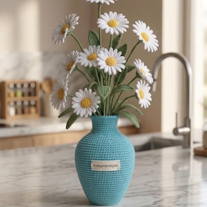 Könnte beinhalten: Ein gehäkeltes Bouquet aus weißen Gänseblümchen mit gelben Zentren und grünen Blättern in einer hellblauen gehäkelten Vase. Die Vase hat ein kleines Etikett mit der Aufschrift "knotgardendigital". Die Anordnung befindet sich auf einer Marmorarbeitsplatte.