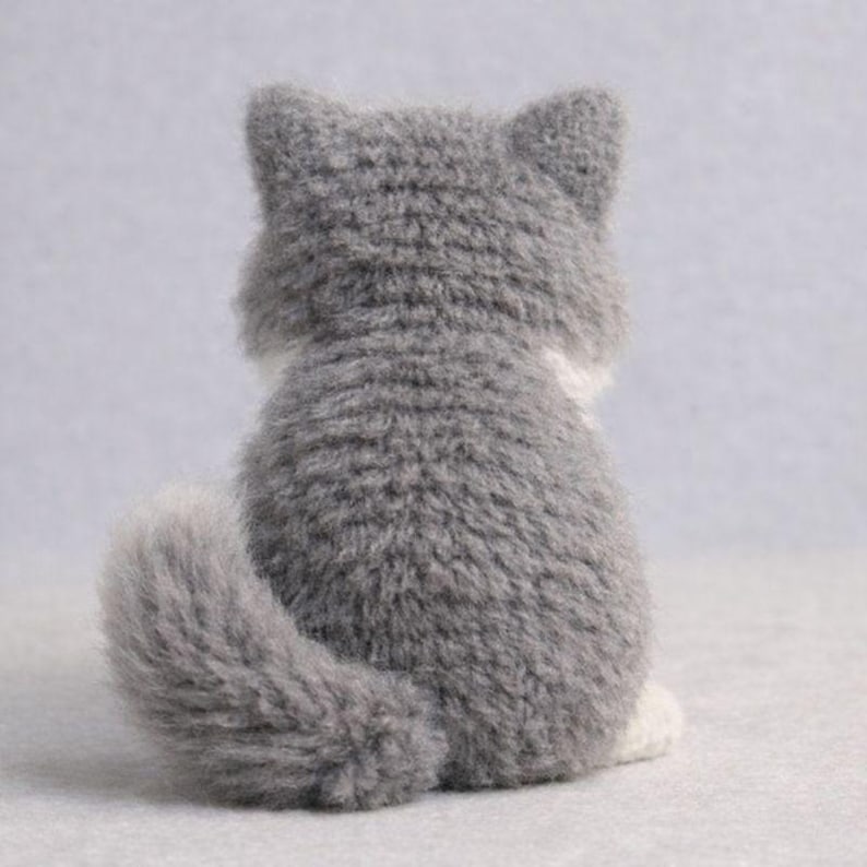 May include: A gray and white plush cat, viewed from the back. The cat has a fluffy tail and ears, with a white patch on its chest. The cat is sitting on a light gray surface.
