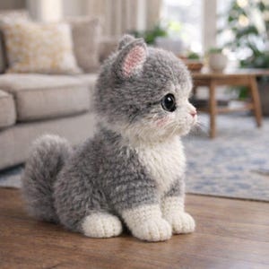 May include: A handmade, gray and white knitted kitten figurine with large black eyes and pink accents. The plush cat has a fluffy tail and is sitting on a wooden surface. The kitten's ears are pink inside, and it has a sweet, innocent expression.
