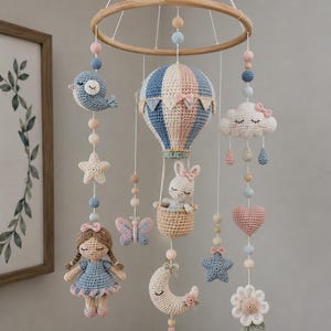 Könnte beinhalten: Handgefertigtes Baby-Mobile aus Häkelarbeit mit Heißluftballon, Häschen, Vogel, Wolke, Sternen, Mond, Blume, Schmetterling, Puppe und Herz. Das Mobile hängt an einem Holzring mit Pastellakzenten. Kinderzimmerdekoration in Blau, Rosa und Creme.