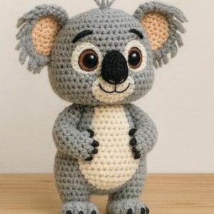 May include: A handmade crochet koala bear in gray and cream colors. The koala has large, expressive eyes, a black nose, and small ears with tan inner details. The toy is standing upright, showcasing its detailed crochet work.