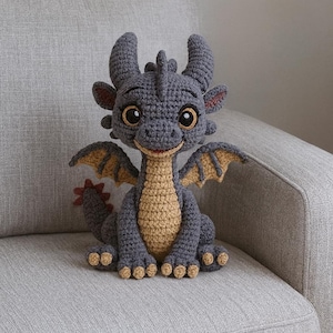 May include: A crocheted dragon figure in grey and tan. The dragon has large eyes, small horns, and detailed wings. It is sitting on a grey sofa, showcasing the handmade craftsmanship of the plush toy.