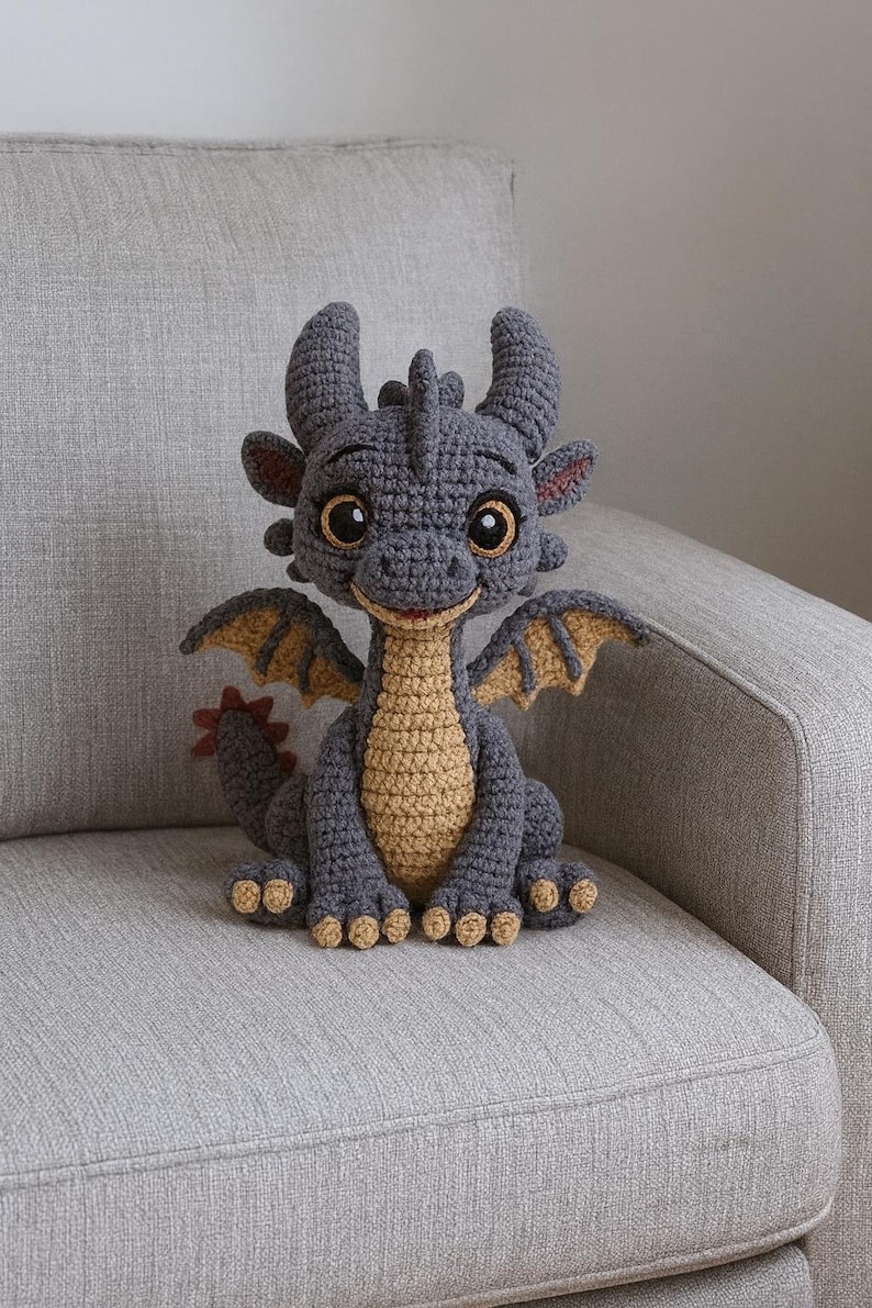 May include: A crocheted dragon figurine in shades of gray and tan. The dragon has large, expressive eyes, small horns, and detailed wings. It is sitting on a gray sofa, showcasing the handmade craftsmanship of the plush toy.
