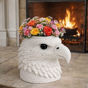 May include: A white crocheted eagle head flower pot filled with colorful roses and other flowers. The eagle has a black eye and a detailed beak. The pot is sitting on a stone floor in front of a fireplace.