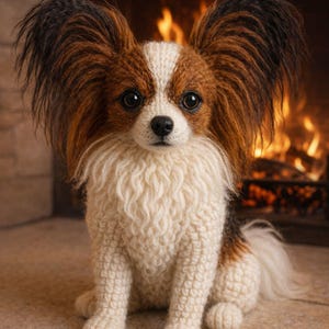 Puede incluir: Escultura artesanal de un perro Papillon hecha de hilo crema, marrón y negro. El perro tiene orejas grandes y con flecos y está sentado frente a una chimenea con un fuego cálido y brillante. Los ojos del perro son oscuros y expresivos.