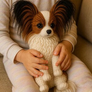 以下が含まれることがあります： 手作りのパピヨン犬のぬいぐるみ。おもちゃは白と茶色の体と、大きくて黒と茶色の耳を持っています。犬は、明るい色の長袖シャツとズボンを着ている人に抱かれています。