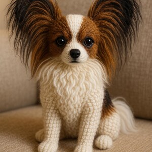 以下が含まれることがあります： 手作りのパピヨン犬の置物。犬は、黒と茶色の毛皮の大きな房状の耳、白と茶色の顔、ふわふわのクリーム色の体を持っています。犬はニュートラルな色の表面に座っています。