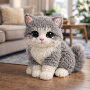 Peut inclure: Un chat en crochet gris et blanc fait à la main, avec de grands yeux noirs, des détails roses sur les oreilles et le nez, et une queue touffue. Le chat est assis sur un sol en bois.