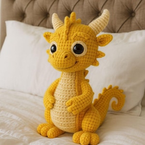 Può includere: Un giocattolo a forma di drago all'uncinetto, giallo e crema, con grandi occhi neri e corna color crema. Il drago sorridente è appoggiato su un letto bianco. Il corpo del drago è fatto di maglia fitta.