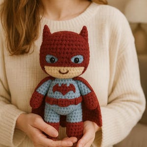 Könnte beinhalten: Handgefertigte Häkelpuppe, die einem Superhelden nachempfunden ist. Die Puppe hat eine rote Kapuze und einen Umhang, einen hellblauen und roten Anzug und ein lächelndes Gesicht mit blauen Augen. Die Puppe ist etwa 15 cm groß.