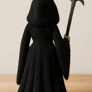 May include: A handmade black crochet Grim Reaper doll, viewed from the back. The doll wears a hooded robe and holds a gray scythe. The scythe's handle is brown, and the doll's hands are tan. The doll is a unique, handcrafted item.