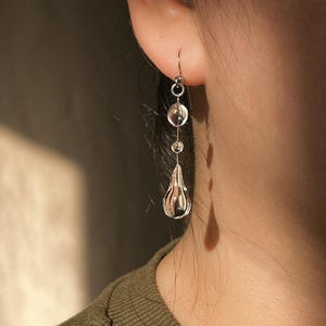 May include: A silver earring with a clear, teardrop-shaped charm. The earring has a hook and features two clear, round beads above the teardrop. The earring is hanging from an ear. The person is wearing a green shirt.