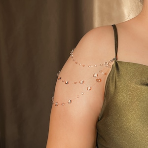 May include: A delicate shoulder chain with multiple strands of small, clear beads and larger, iridescent, circular beads. The jewelry is worn over the shoulder and arm, creating a layered effect. The background is a solid, neutral color.