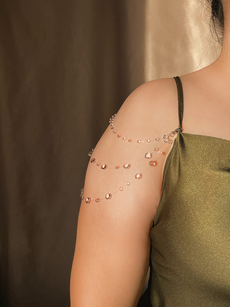 May include: A delicate shoulder chain with multiple strands of small, clear beads and larger, iridescent, circular beads. The jewelry is worn over the shoulder and arm, creating a layered effect. The background is a solid, neutral color.