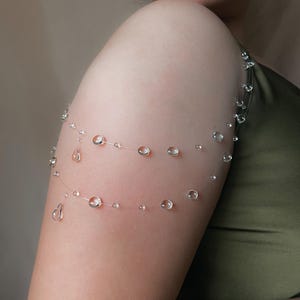 K&ouml;nnte beinhalten: Zarter Armschmuck mit klaren, runden und tropfenf&ouml;rmigen Perlen, die auf einem d&uuml;nnen, transparenten Draht aufgereiht sind. Die Perlen sind in zwei parallelen Linien um den Oberarm angeordnet und bilden ein subtiles, elegantes Accessoire.