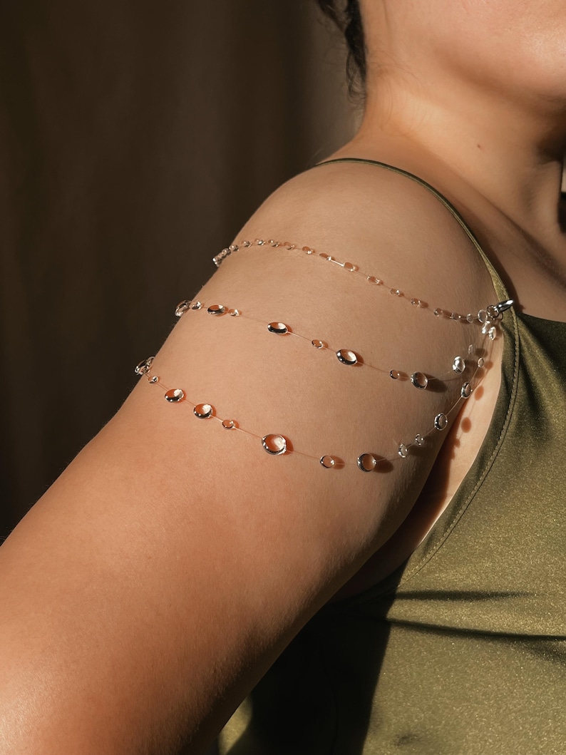May include: Delicate shoulder jewelry featuring three strands of clear, thin chain adorned with clear and amber-colored beads. The jewelry is clipped to the shoulder and arm, creating a unique and elegant accessory.