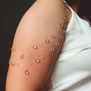 K&ouml;nnte beinhalten: Eine zarte Armkette mit klaren, runden Perlen und tropfenf&ouml;rmigen Anh&auml;ngern. Der Schmuck ist auf einem fast unsichtbaren Faden aufgef&auml;delt, wodurch ein schwebender Effekt entsteht. Die Perlen sind in verschiedenen Gr&ouml;&szlig;en gehalten, was diesem einzigartigen Accessoire einen visuellen Reiz verleiht.