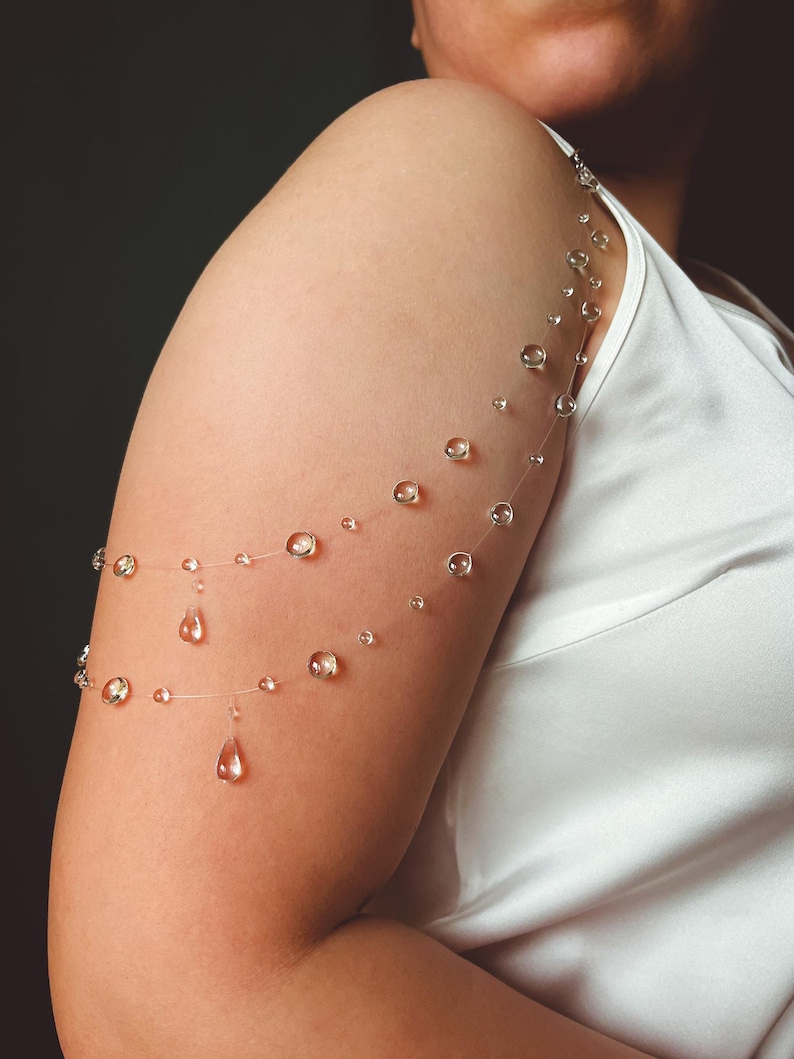 K&ouml;nnte beinhalten: Eine zarte Armkette mit klaren, runden Perlen und tropfenf&ouml;rmigen Anh&auml;ngern. Der Schmuck ist auf einem fast unsichtbaren Faden aufgef&auml;delt, wodurch ein schwebender Effekt entsteht. Die Perlen sind in verschiedenen Gr&ouml;&szlig;en gehalten, was diesem einzigartigen Accessoire einen visuellen Reiz verleiht.