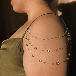 May include: A delicate shoulder necklace featuring three strands of clear beading with iridescent, copper-colored accents. The necklace is attached to a small, silver-toned clasp at the shoulder.