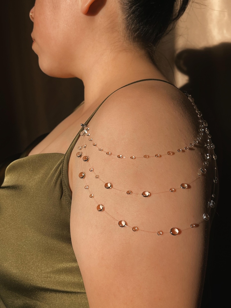 May include: A delicate shoulder necklace featuring three strands of clear beading with iridescent, copper-colored accents. The necklace is attached to a small, silver-toned clasp at the shoulder.