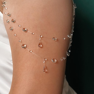 Puede incluir: Una delicada cadena de brazo transparente con múltiples cuentas redondas y transparentes y colgantes en forma de lágrima. La joya está ensartada en un fino hilo transparente, creando un efecto flotante. La cadena de brazo se lleva en la parte superior del brazo.