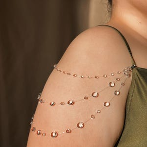 May include: A delicate, layered shoulder jewelry piece. Three strands of clear, beaded strands cascade across the shoulder and arm. The beads are clear and round, with a subtle, reflective quality. The jewelry is attached with a small clasp.