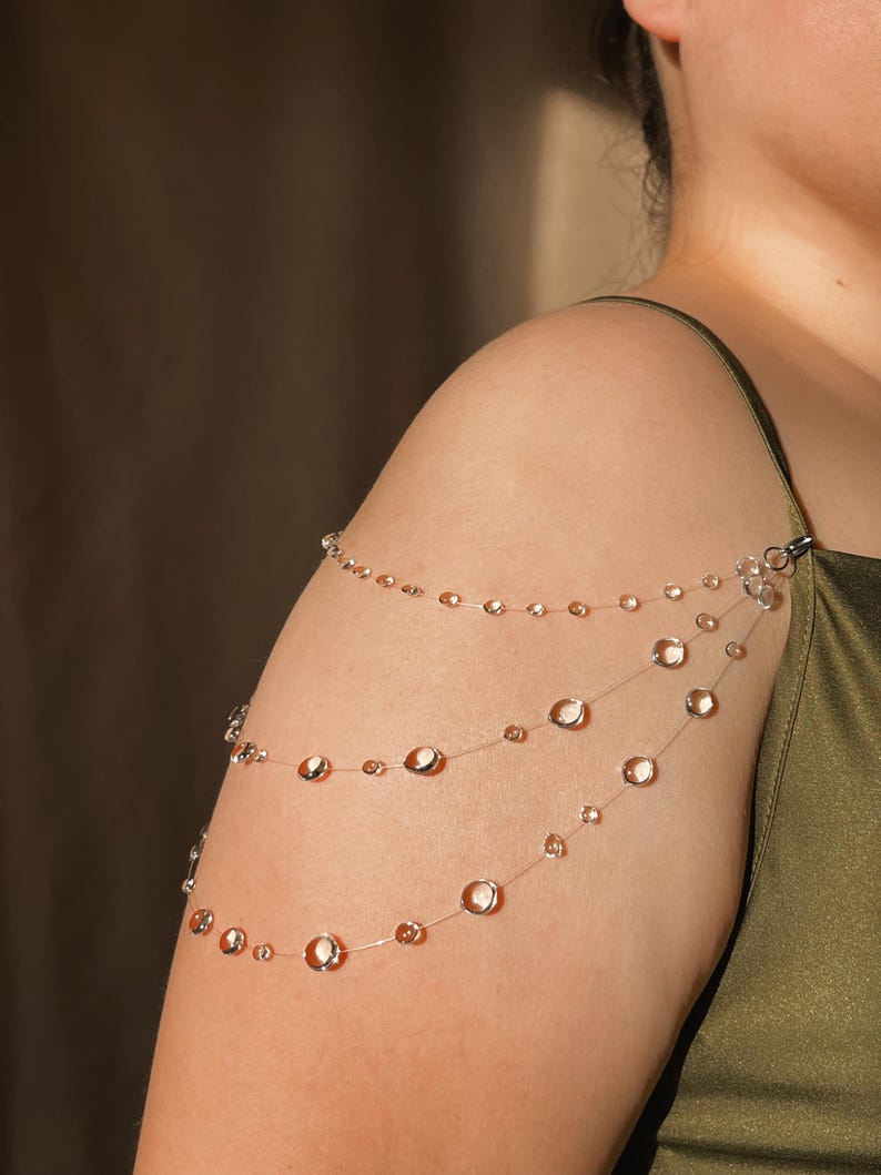 May include: A delicate, layered shoulder jewelry piece. Three strands of clear, beaded strands cascade across the shoulder and arm. The beads are clear and round, with a subtle, reflective quality. The jewelry is attached with a small clasp.