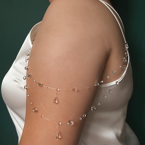 K&ouml;nnte beinhalten: Eine transparente Armkette mit tropfenf&ouml;rmigen und runden Kristallperlen. Der Schmuck ist auf einem d&uuml;nnen, transparenten Draht aufgef&auml;delt, wodurch ein Schwebeeffekt entsteht. Die Armkette wird &uuml;ber einem wei&szlig;en Satin-Top getragen, mit einem dunkelgr&uuml;nen Hintergrund.