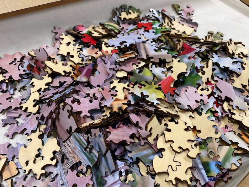 May include: A pile of wooden jigsaw puzzle pieces with a variety of colors and images. The pieces are scattered on a white surface.