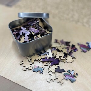 May include: A metal tin filled with wooden puzzle pieces. The pieces are a mix of light brown and purple. Some of the pieces are scattered on a light brown surface.