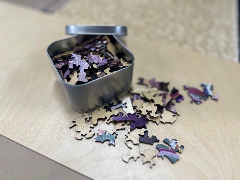 May include: A metal tin filled with wooden puzzle pieces. The pieces are a mix of light brown and purple. Some of the pieces are scattered on a light brown surface.