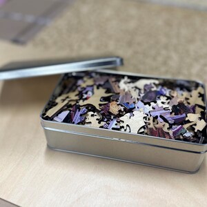 May include: A silver metal box with a lid open, revealing a jumbled pile of wooden jigsaw puzzle pieces. The pieces are various shapes and sizes, with a colorful printed image on the front side.