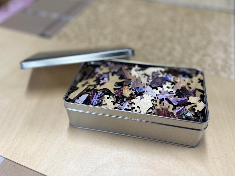 May include: A silver metal box with a lid open, revealing a jumbled pile of wooden jigsaw puzzle pieces. The pieces are various shapes and sizes, with a colorful printed image on the front side.