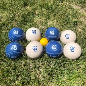 May include: Eight bocce balls, seven with a white and blue design and the text "GB" and one yellow ball. The balls are arranged in a circle on a green grassy surface.