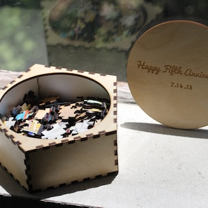 May include: A wooden box with a round lid. The lid is engraved with the text "Happy Fifth Anniversary 7.14.13". The box is filled with puzzle pieces.