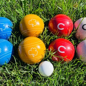 Op de afbeelding: Een set jeu de boules ballen in groen gras. De set bevat blauwe ballen met witte klavers, gele ballen met witte strepen en klavers, rode ballen met een witte "C" en witte ballen met een zwarte "C".